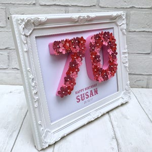 May include: A white framed picture with a pink background and a 3D number 70 made of pink and red flowers. The text "Happy Birthday Susan 29.10.17" is printed below the number.