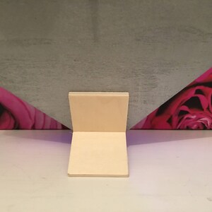 Pode incluir: Um suporte de cart&atilde;o branco com uma base quadrada e dois lados retos. O suporte est&aacute; posicionado em frente a um fundo com um padr&atilde;o floral rosa e roxo.
