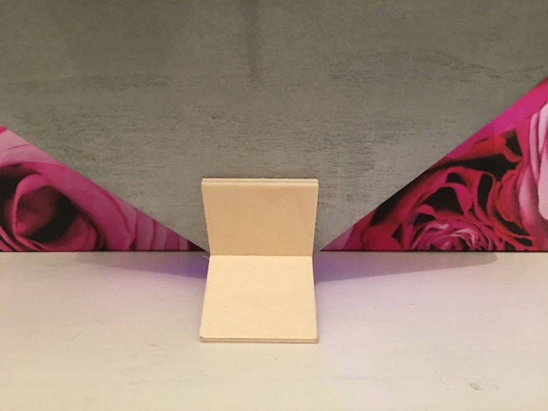 Pode incluir: Um suporte de cart&atilde;o branco com uma base quadrada e dois lados retos. O suporte est&aacute; posicionado em frente a um fundo com um padr&atilde;o floral rosa e roxo.