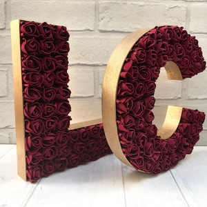 May include: Two large, gold-framed letters, "L" and "C", are covered in burgundy red fabric roses. The letters are freestanding and would be ideal for a wedding or party decoration.