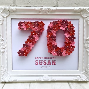 May include: A white framed picture with a pink background. The number 70 is made of pink and red flowers. The text "Happy Birthday Susan 29.10.17" is below the number.