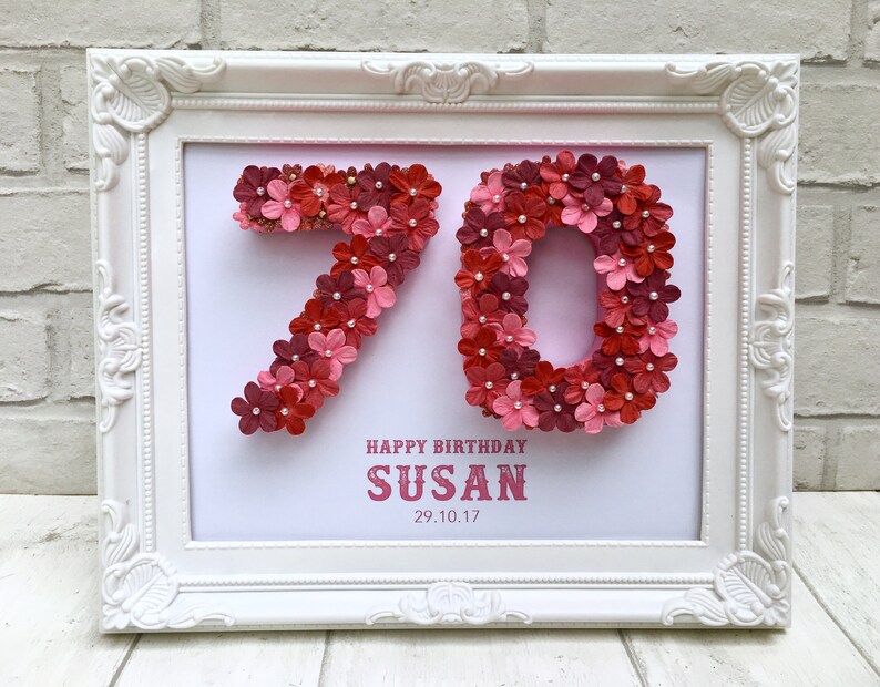 May include: A white framed picture with a pink background. The number 70 is made of pink and red flowers. The text "Happy Birthday Susan 29.10.17" is below the number.