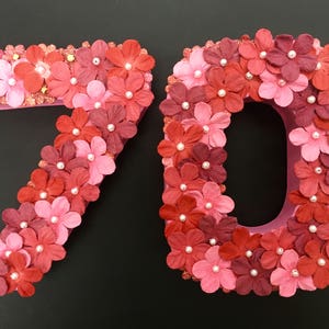 May include: Two large numbers, 7 and 0, made of pink cardboard and covered in red and pink paper flowers. The flowers have white pearl centres.