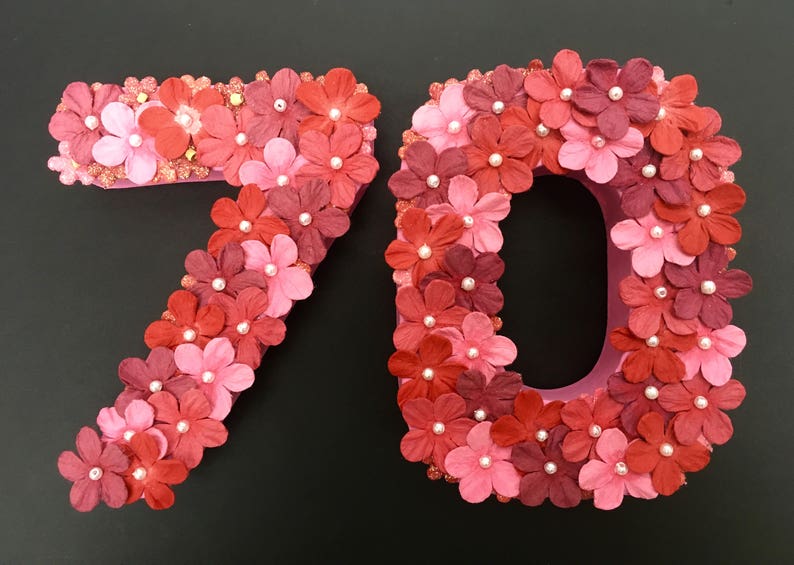 May include: Two large numbers, 7 and 0, made of pink cardboard and covered in red and pink paper flowers. The flowers have white pearl centres.