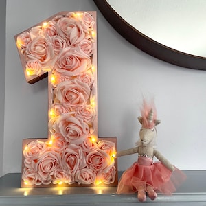 Puede incluir: Un número uno iluminado de color rosa y dorado hecho de rosas de tela. El número uno está sobre una superficie gris con una muñeca de unicornio blanca delante.