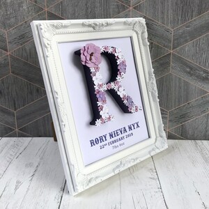 May include: White framed picture with a black letter R decorated with purple and white flowers. The text below the letter reads "Rory Nieva Nyx 22nd February 2019 7lbs 6oz"