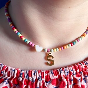 Peut inclure: Un collier de perles colorées avec un pendentif en forme de cœur blanc et une lettre 'S' dorée.