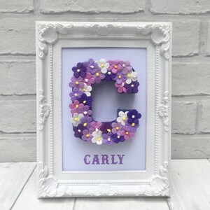 May include: White frame with a purple and white letter C made of flowers. The name Carly is written below the letter.