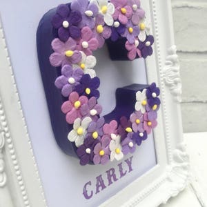 May include: A white frame with a purple background and a purple letter C decorated with purple, pink, and white flowers. The name "CARLY" is printed below the letter.