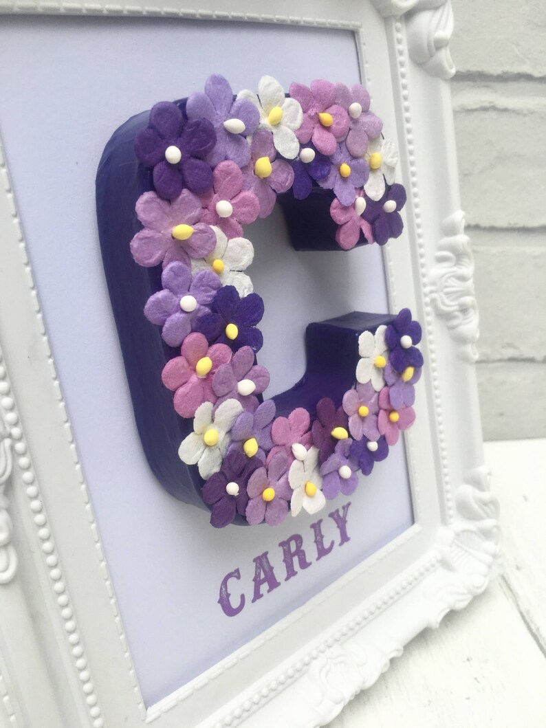 May include: A white frame with a purple background and a purple letter C decorated with purple, pink, and white flowers. The name "CARLY" is printed below the letter.