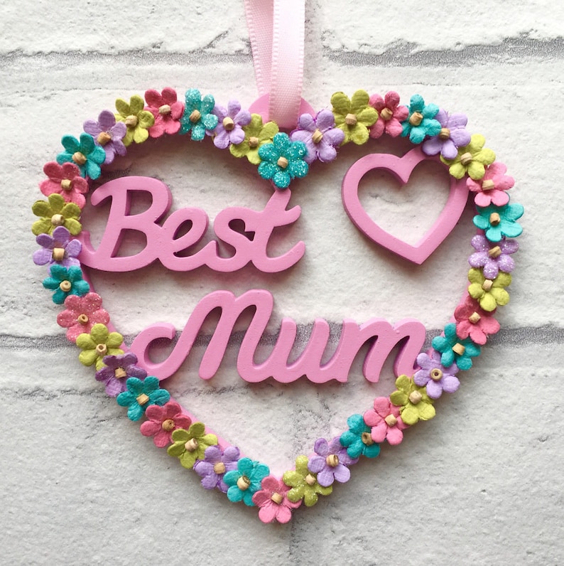 May include: Pink wooden heart-shaped sign with the words "Best Mum" in white lettering. The heart is decorated with a border of colorful paper flowers.