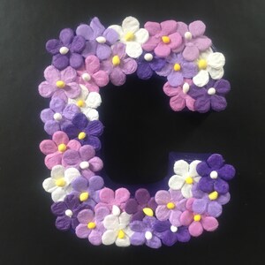May include: A purple letter C decorated with white, pink, and purple paper flowers. The flowers have yellow centers.