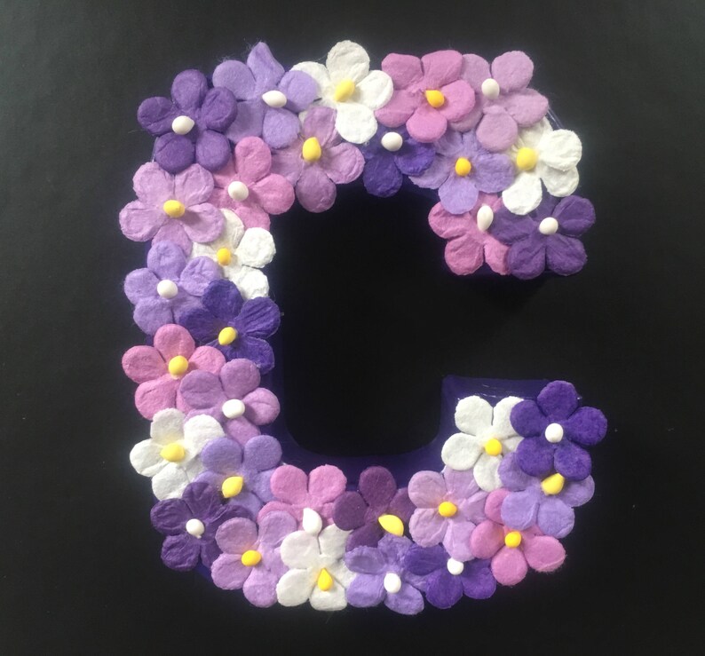 May include: A purple letter C decorated with white, pink, and purple paper flowers. The flowers have yellow centers.