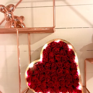 Pode incluir: Um quadro de madeira em forma de cora&ccedil;&atilde;o cheio de rosas vermelhas e iluminado com luzes brancas quentes. O quadro est&aacute; sentado em uma prateleira de metal rosa dourado.