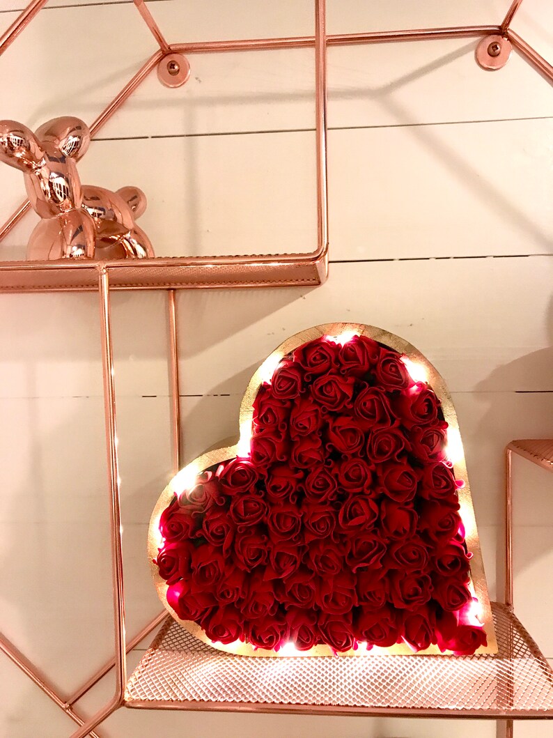 Pode incluir: Um quadro de madeira em forma de cora&ccedil;&atilde;o cheio de rosas vermelhas e iluminado com luzes brancas quentes. O quadro est&aacute; sentado em uma prateleira de metal rosa dourado.