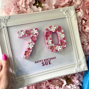 May include: A white framed picture with a pink background. The numbers 70 are made of pink and white flowers. The text "Happy Birthday Sue" is written in red below the numbers.