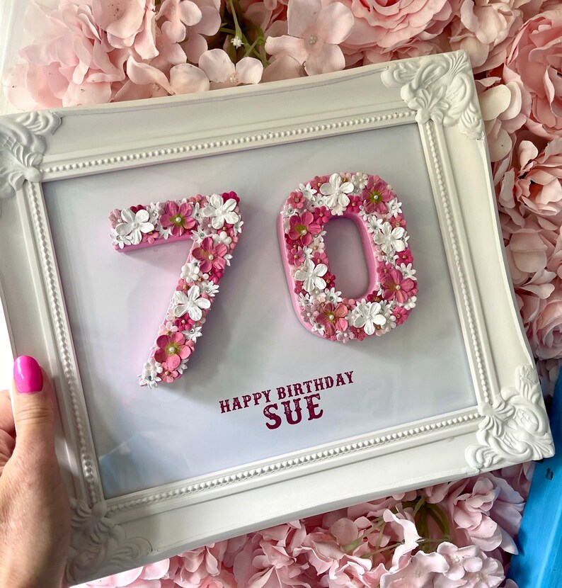 May include: A white framed picture with a pink background. The numbers 70 are made of pink and white flowers. The text "Happy Birthday Sue" is written in red below the numbers.