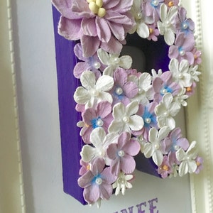 May include: A purple letter R decorated with white and lavender paper flowers and pearl accents. The letter is framed and the name "RENEE" is written below the letter in purple.