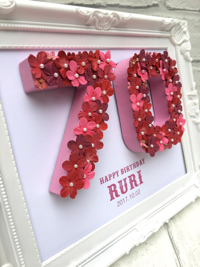 May include: A white frame with a pink background and the numbers 70 made of pink paper flowers. The flowers are decorated with small white pearls. The text "Happy Birthday RURI 2017.10.02" is printed in red below the numbers.