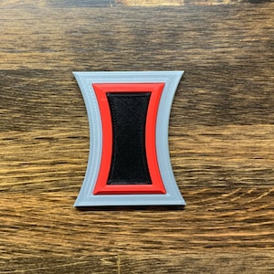 May include: A gray, red, and black 3D printed emblem shaped like an hourglass. The emblem has a black center and a red outline.