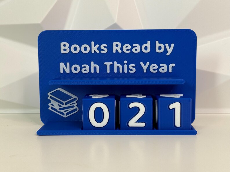 Może przedstawiać: Niebieskie akcesorium biurowe z napisem "Books Read by Noah This Year". Posiada p&oacute;łkę z grafiką stosu książek i trzy obracające się kostki wyświetlające liczbę 021. Akcesorium służy do śledzenia postęp&oacute;w w czytaniu.