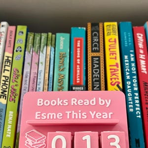 Może przedstawiać: R&oacute;żowy stojak na książki z napisem "Books Read by Esme This Year" i numerem "013". Stojak stoi przed p&oacute;łką z kolorowymi książkami.