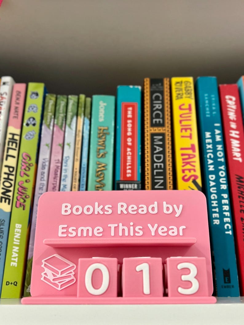 Może przedstawiać: R&oacute;żowy stojak na książki z napisem "Books Read by Esme This Year" i numerem "013". Stojak stoi przed p&oacute;łką z kolorowymi książkami.