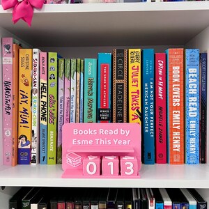Może przedstawiać: Biały regał prezentuje kolekcję książek z kolorowymi grzbietami. R&oacute;żowy znak głosi "Books Read by Esme This Year" z licznikiem pokazującym "013". Ozdoby, takie jak r&oacute;żowa kokarda, znajdują się na p&oacute;łce.