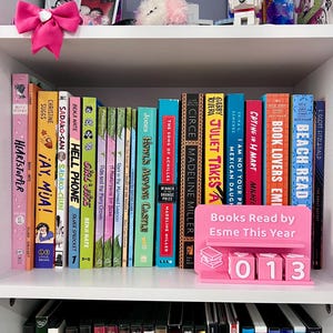 Może przedstawiać: Biały regał prezentuje kolekcję książek z kolorowymi grzbietami. R&oacute;żowy stojak na książki głosi "Books Read by Esme This Year" z numerem "013". R&oacute;żowa kokarda i elementy dekoracyjne znajdują się na g&oacute;rnej p&oacute;łce.