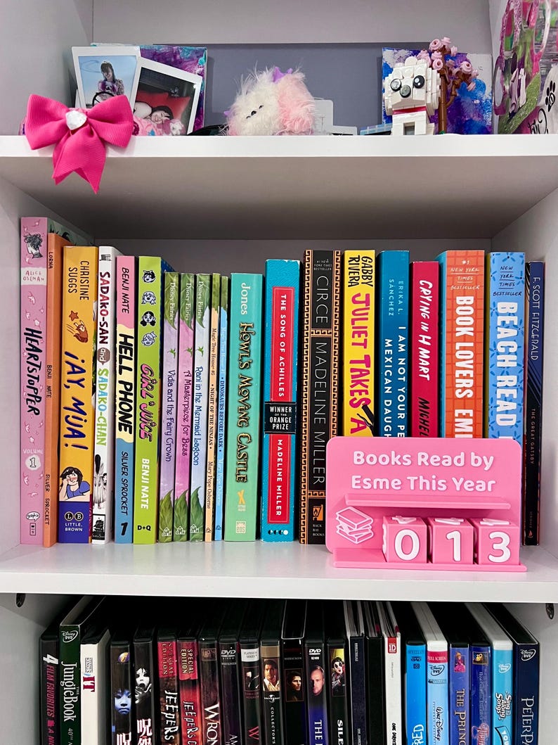 Może przedstawiać: Biały regał prezentuje kolekcję książek z kolorowymi grzbietami. R&oacute;żowy stojak na książki głosi "Books Read by Esme This Year" z numerem "013". R&oacute;żowa kokarda i elementy dekoracyjne znajdują się na g&oacute;rnej p&oacute;łce.