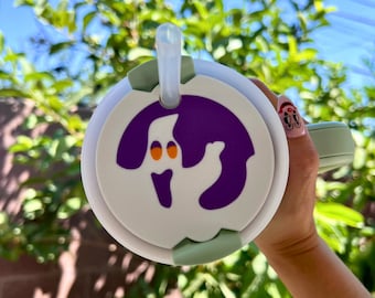 May include: White reusable cup with a green lid and a purple ghost design. The cup has a clear straw sticking out of the lid.