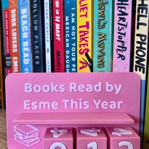 Może przedstawiać: R&oacute;żowy, drukowany w 3D stojak na książki z napisem "Books Read by Esme This Year". Stojak ma p&oacute;łkę i trzy obracające się kostki z numerami pokazującymi "013". W tle kolekcja kolorowych książek.
