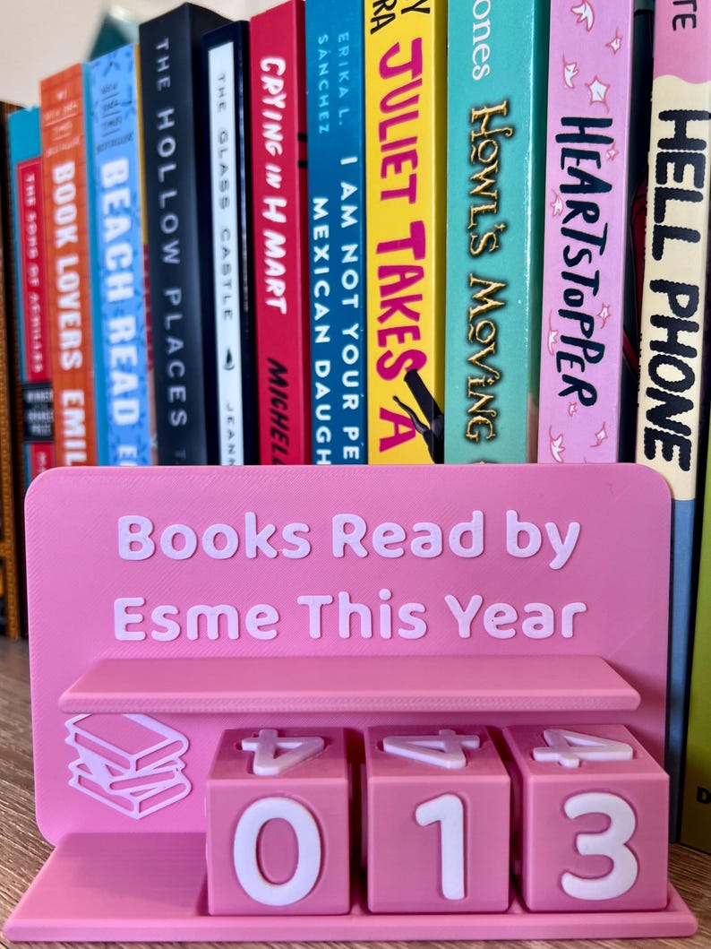 Może przedstawiać: R&oacute;żowy, drukowany w 3D stojak na książki z napisem "Books Read by Esme This Year". Stojak ma p&oacute;łkę i trzy obracające się kostki z numerami pokazującymi "013". W tle kolekcja kolorowych książek.