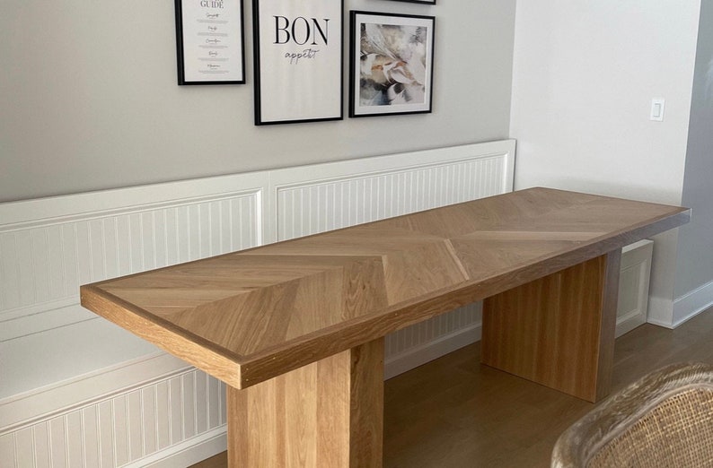 May include: A long, wooden table with a chevron pattern top and two rectangular legs. The table is made of light wood and has a natural finish.