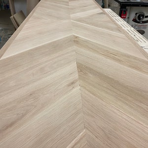 May include: A close-up view of a light wood chevron pattern floor. The wood is a light beige color and the chevron pattern is made up of narrow strips of wood.