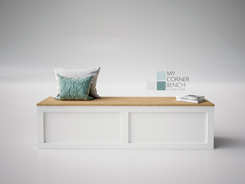 May include: A white bench with a light wood top, featuring two decorative cushions in shades of grey and teal. The bench has a rectangular shape and the text "MY CORNER BENCH CUSTOM MADE" is visible.