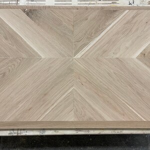 May include: A close-up of a light brown wood floor with a herringbone pattern. The wood is arranged in a geometric pattern with alternating light and dark stripes.