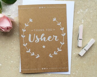 May include: A brown card with a white wreath and the text "Thank You Usher" inside. The card is on a white background with pink and white patterned clothespins.