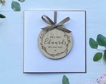 May include: A white card with a brown wooden circle ornament attached. The ornament has a floral design and the text "MR & MRS Edwards 28TH MAY 2023".