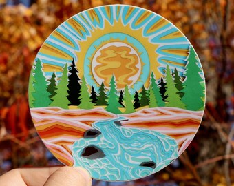 May include: A round sticker with a psychedelic design featuring a sun, trees, and a river. The sun is yellow and orange with a swirling pattern. The trees are green and the river is blue with white swirls. The background is a mix of orange, pink, and yellow.
