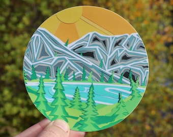 May include: A circular sticker featuring a layered paper-cut style landscape illustration. The design depicts a bright yellow sun over jagged gray and white mountains, a light blue lake, and several vibrant green pine trees in the foreground.