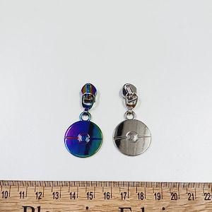 以下が含まれることがあります： 銀色と虹色の仕上げのジッパーのプルが2つ。プルは、赤と白のデザインの丸いオブジェクトであるポケボールの形をしています。プルは、木製の定規のある白い背景に対して表示されています。