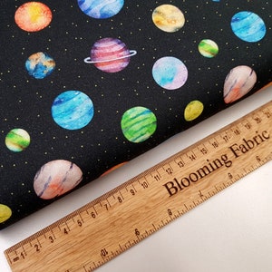 May include: Black fabric with a repeating pattern of colorful planets on a black background. The fabric is measured with a wooden ruler that says "Blooming Fabric".