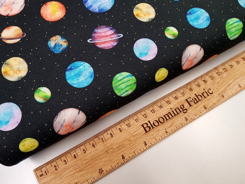 May include: Black fabric with a repeating pattern of colorful planets on a black background. The fabric is measured with a wooden ruler that says "Blooming Fabric".