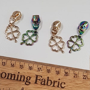 May include: Four gold-coloured zipper pulls with a four-leaf clover charm. The charms are iridescent with a rainbow effect. The zipper pulls are shown on a wooden ruler with the text "Coming Fabric" printed on it.