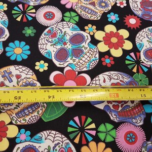 May include: Black fabric with a repeating pattern of colorful sugar skulls and flowers. The fabric is measured with a yellow ruler.