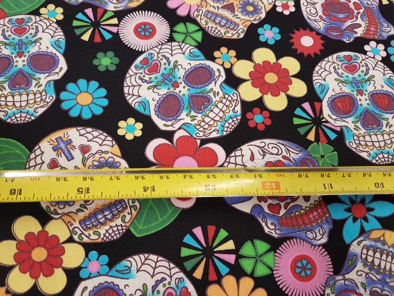 May include: Black fabric with a repeating pattern of colorful sugar skulls and flowers. The fabric is measured with a yellow ruler.