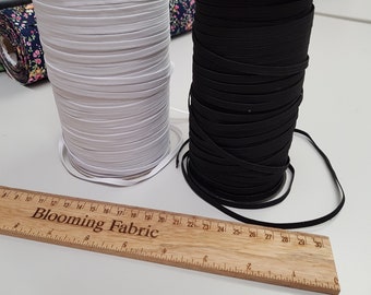 Pode incluir: Dois rolos de cordão elástico, um branco e outro preto, são exibidos em uma superfície branca. Uma régua de madeira com as palavras "Blooming Fabric" está em primeiro plano.