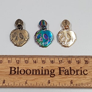 May include: Three zipper pulls in different finishes: gold, antique brass, and iridescent. Each pull features a heart-shaped design with a skull and crossbones inside.