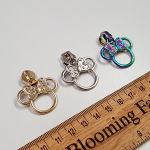 Może przedstawiać: Trzy suwaki do zamka błyskawicznego w kształcie głowy myszy z kreskówek z kokardką. Suwaki są srebrne, złote i tęczowe.